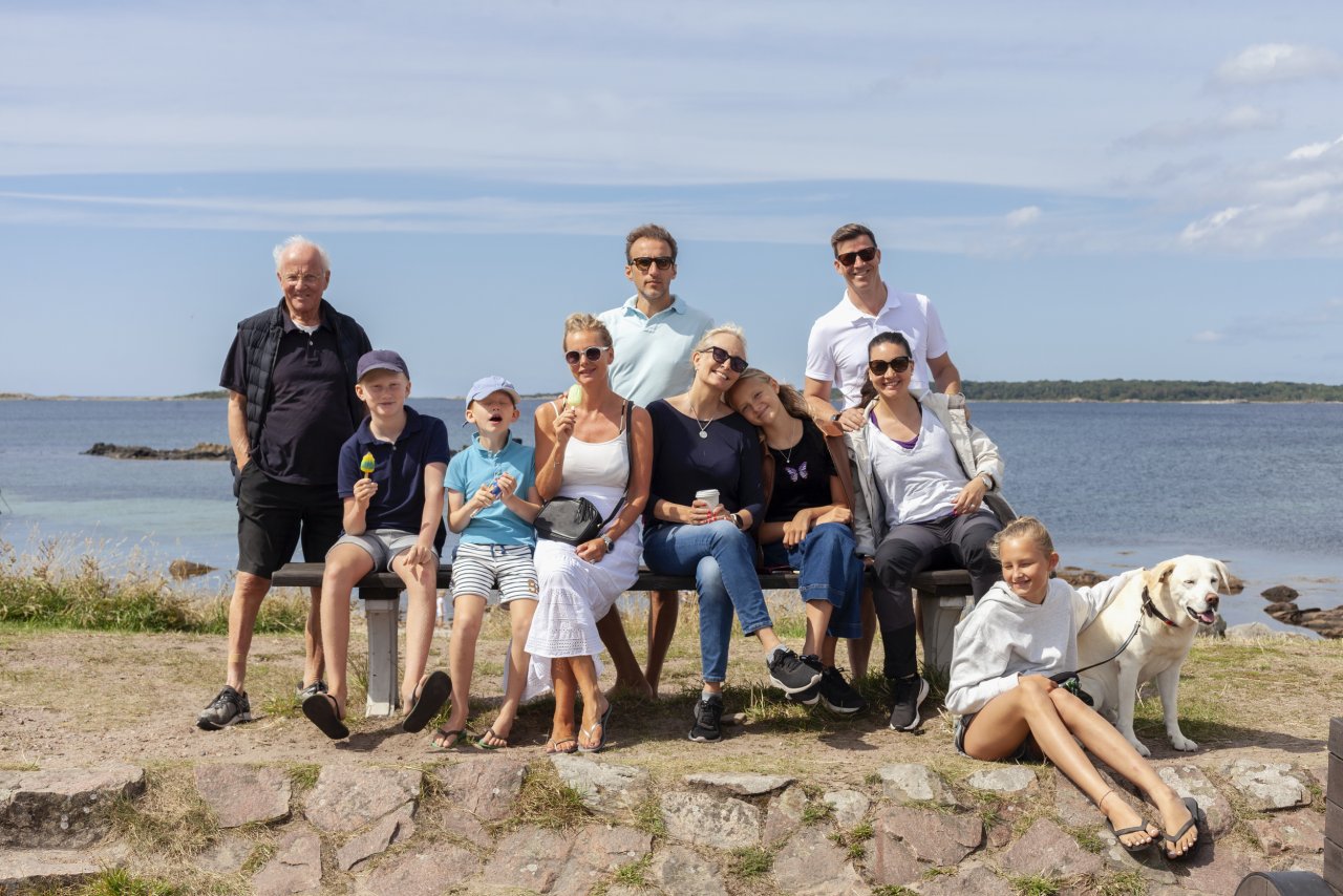Stor grupp med familj sitter på bänk vid havet en fin sommardag.