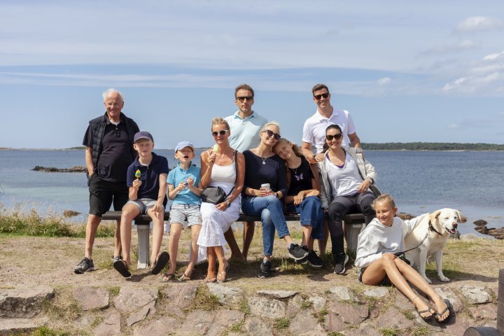 Stor grupp med familj sitter på bänk vid havet en fin sommardag.