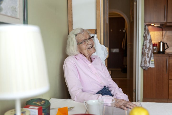 Äldre , glad kvinna sitter vid sitt köksbord.