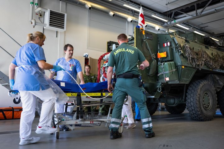 Sjukvardsövning med militärer och sjukvårdsperonal. De är samlade runt en skadad person på en bår och ska lastas in i ett militärfordon,