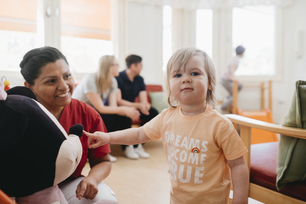 Sköterska och liten tjej på familjecentralen med familj i bakgrunden