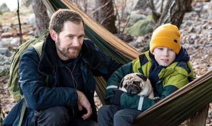 Man sitter bredvid en pojke med hund i en hängmatta ute i naturen