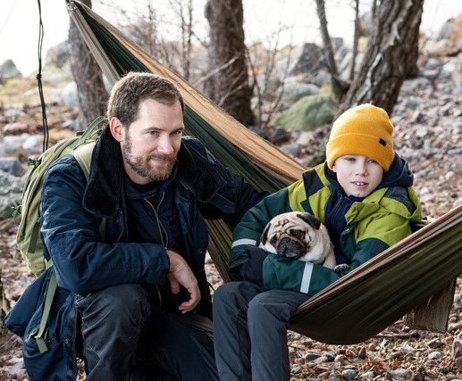 Man sitter bredvid en pojke med hund i en hängmatta ute i naturen